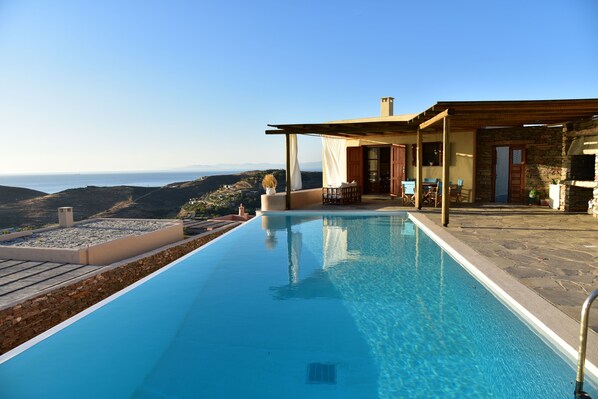 Ein Pool mit freiem Blick auf den Ozean, eine überdachte Terrasse und ein Steinhaus mit Schornstein.