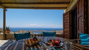 Un tavolo apparecchiato con frutta, una ciotola di yogurt e una tazza, con vista panoramica sul mare e le colline.