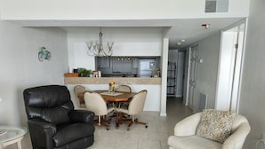 A living room with a leather chair, a glass coffee table, and a patterned rug. There is a dining area with a table and chairs, and a kitchen in the background. - Oceanfront - Beautiful, Direct Oceanfront. Extra Large 1 Bedroom, 1+ Bath (New Smyrna Beach)