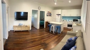A modern living room with a wooden floor, a sofa, a TV, and a kitchen area with white cabinets and a blue countertop. - A beach house with colour! (Shoal Bay)