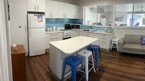 A modern kitchen with white cabinets, a central island, blue stools, and a view through the window. - A beach house with colour! (Shoal Bay)