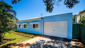 A single-story house with a blue exterior, a white garage door, and a small front yard with a brick pathway and a garden bed. - A beach house with colour! (Shoal Bay)
