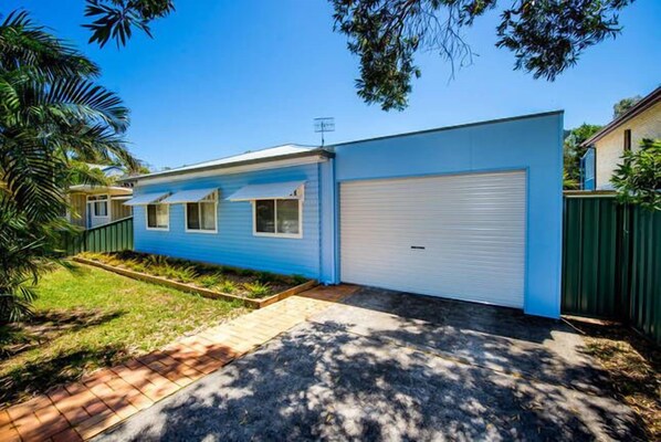A single-story house with a blue exterior, a white garage door, and a small front yard with a brick pathway and a garden bed. - A beach house with colour! (Shoal Bay)