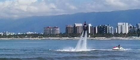 A waterfront cityscape with a fountain, a jet ski, and a distant mountain range.