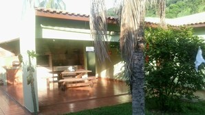 Ein überdachter Patio-Bereich mit Holztisch und Bänken, umgeben von viel Grün und einer Palme.