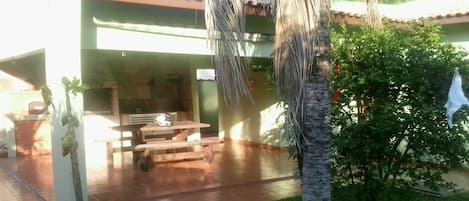 Un patio techado con mesa y bancos de madera, rodeado de follaje y una palmera.