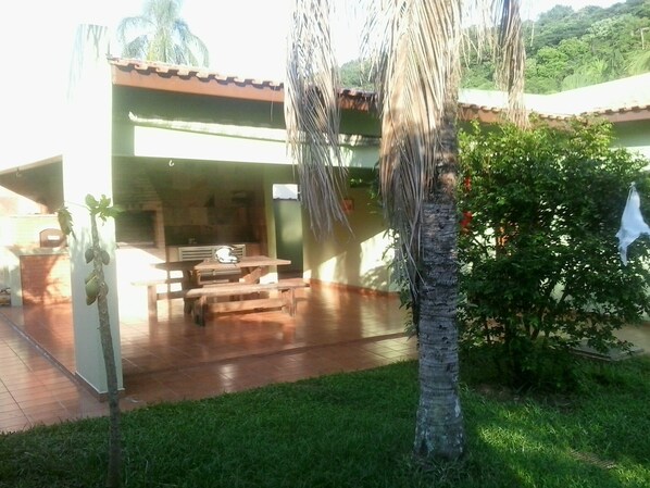 A covered patio area with a wooden table and benches, surrounded by greenery and a palm tree. - House with air conditioning 50m from Riviera Beach, house with Wi-Fi.  (Bertioga)