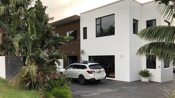 Ein modernes, zweistöckiges Haus mit weißer Fassade, ein weißes Auto auf der Auffahrt und üppige tropische Pflanzen rund um das Grundstück.