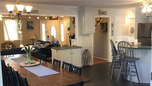 Un grand espace dédié à la salle à manger, avec une longue table en bois et des chaises, un coin cuisine équipé d’un réfrigérateur et de tabourets de bar, et un salon agrémenté d’un canapé et d’objets décoratifs.