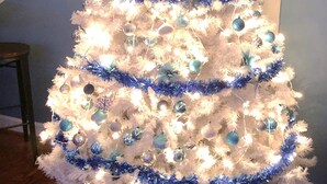 Un sapin de Noël blanc, décoré d’ornements bleus et de guirlandes lumineuses, est placé dans une pièce qui comprend un escalier et un mur décoratif.