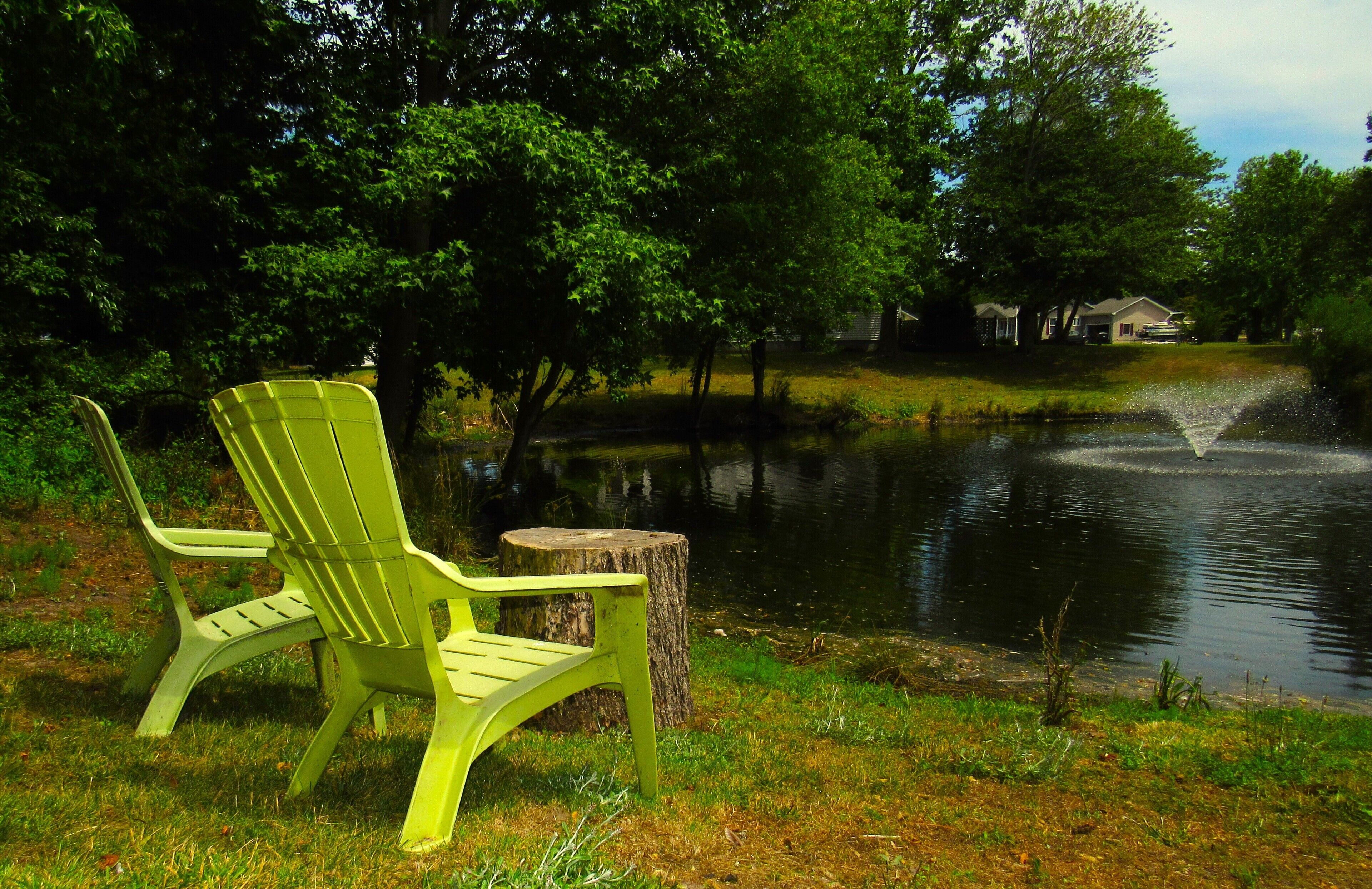 A green Adirondack chair by a pond with a wooden stump nearby.