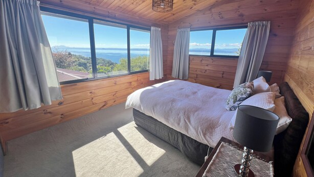 A wooden cabin bedroom with a large bed, a nightstand, a lamp, and a view of the landscape through large windows. - Lake views, boating, skiing and hot pools (Omori)