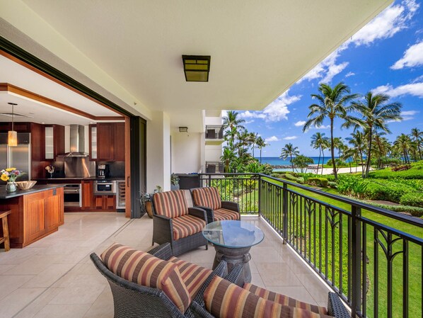 A balcony with wicker furniture and a glass table, overlooking a beach with palm trees and the ocean. - Nalu Collection 2/2 Oceanfront Living~Luxurious Amenities (Kapolei)