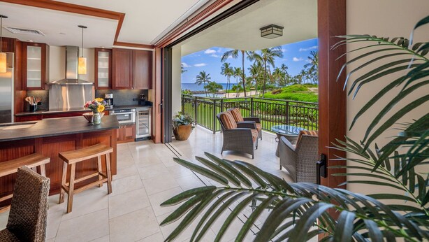 A modern kitchen with a large island, a dining area with wicker furniture, and a balcony with a view of the ocean and palm trees. - Nalu Collection 2/2 Oceanfront Living~Luxurious Amenities (Kapolei)