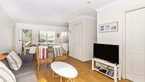 A modern living room with a sofa, a dining table with chairs, a TV on a stand, and a kitchen in the background. - Horizons in Mollymook (Mollymook Beach)