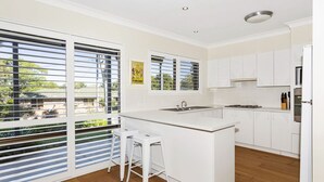 A modern kitchen with white cabinetry, a central island, and large windows with blinds. - Horizons in Mollymook (Mollymook Beach)