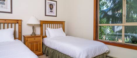 Una camera da letto con due letti singoli, un comodino con una lampada, quadri appesi alle pareti e un'ampia finestra che dà su alberi e palazzi.