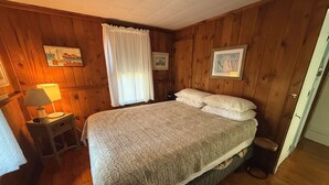 A wooden-paneled room with a bed, a nightstand with a lamp, and a door.