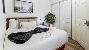 Een net opgemaakt bed met witte lakens, een zwarte plaid, een bedlampje en een plantje op een nachtkastje.