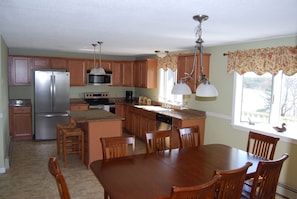 A kitchen with wooden cabinets, a stainless steel refrigerator, a microwave, and a dining area with a wooden table and chairs. - Charming home on Montsweag Bay.  (Woolwich)