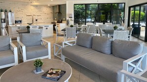 Uma sala de estar moderna com sofá, poltronas e uma mesa de centro. Ao fundo, cozinha com bancada branca e eletrodomésticos de aço inoxidável.