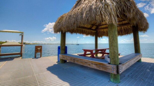 Um deck de madeira com um gazebo de telhado de palha, uma mesa de piquenique e um banco.