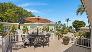 Une terrasse en bois avec une table et des chaises, entourée de clôtures blanches en bois et de plantes tropicales.