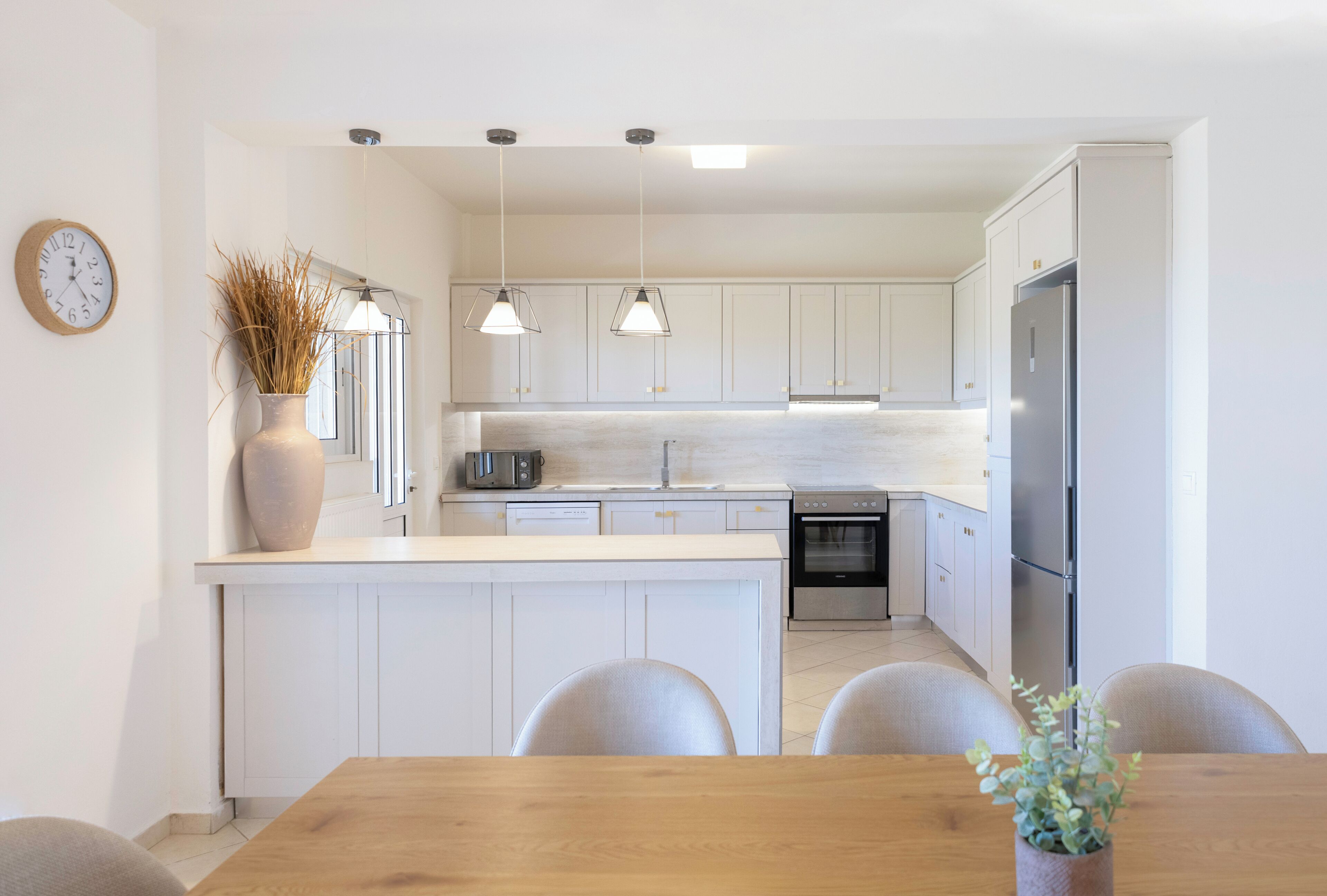 A modern kitchen with white cabinets, a wooden dining table, and pendant lights.