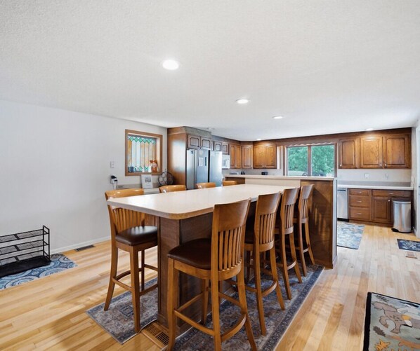 A modern kitchen with wooden cabinets, a central island with chairs, and a large window providing natural light.