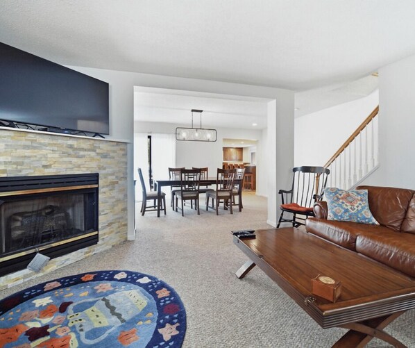 A modern living room with a fireplace, a dining area, and a staircase.