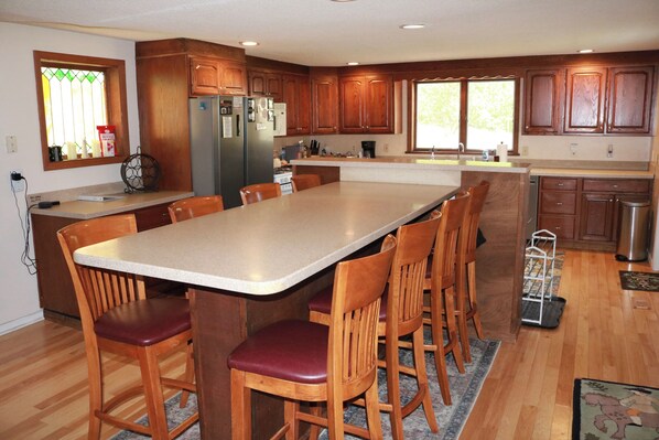 A kitchen with wooden cabinets, a central island with chairs, a refrigerator, and a window with a view.