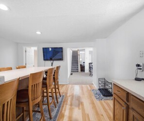 A modern kitchen with a dining area, wooden chairs, a flat-screen TV, and a staircase leading to another level.