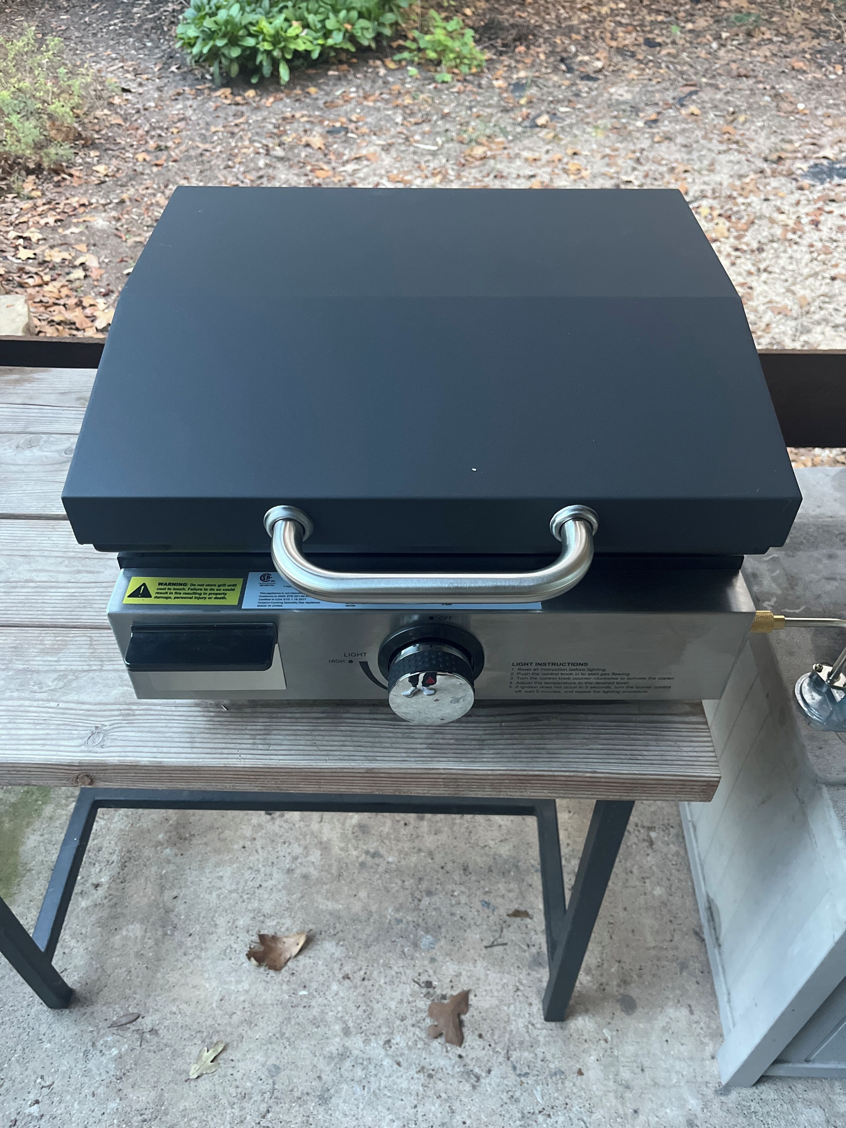 A black portable grill with a metal handle and a control knob, placed on a metal table outdoors.