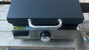 A black portable grill with a metal handle and a control knob, placed on a metal table outdoors.