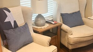 Une pièce avec deux fauteuils beiges, dont l’un est agrémenté d’un coussin en forme d’étoile, une petite table avec une lampe et une fenêtre avec des stores.
