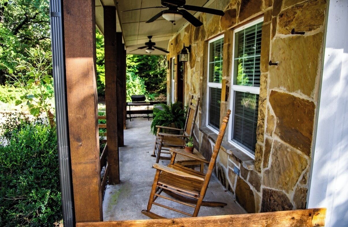 A covered porch with stone walls, wooden chairs, and a ceiling fan.