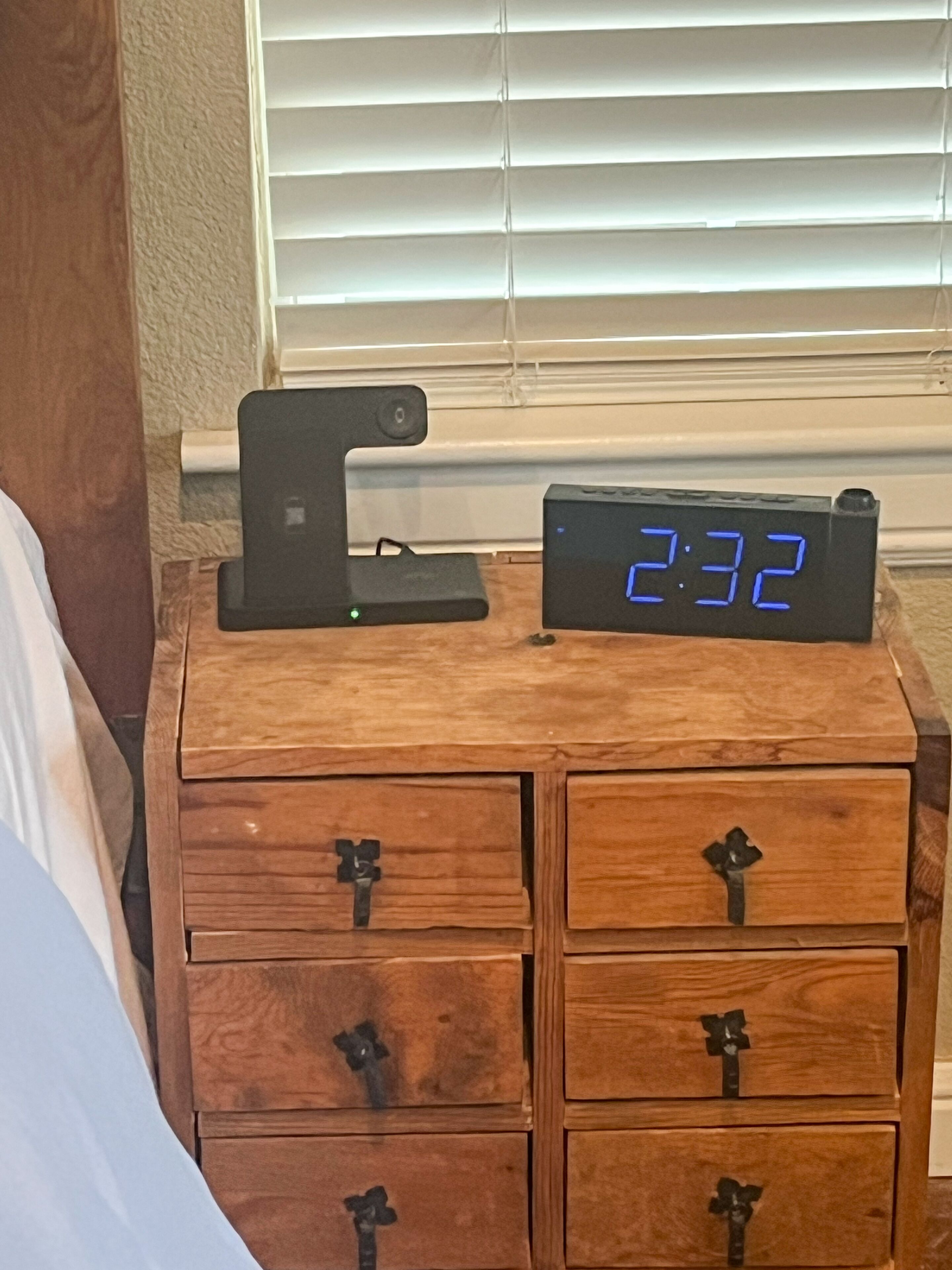 A wooden nightstand with a digital clock displaying 7:32, a smartphone charging dock, and multiple drawers with metal handles.