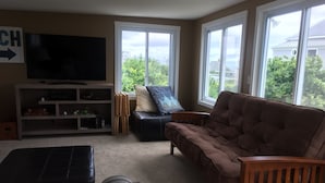 Ein Wohnzimmer mit einem braunen Sofa, einem schwarzen Ledersessel und einem gestreiften Hocker. Ein Fernseher steht auf einem Lowboard, ein Holzstuhl und ein Fenster mit Blick auf Grünflächen sind ebenfalls vorhanden.
