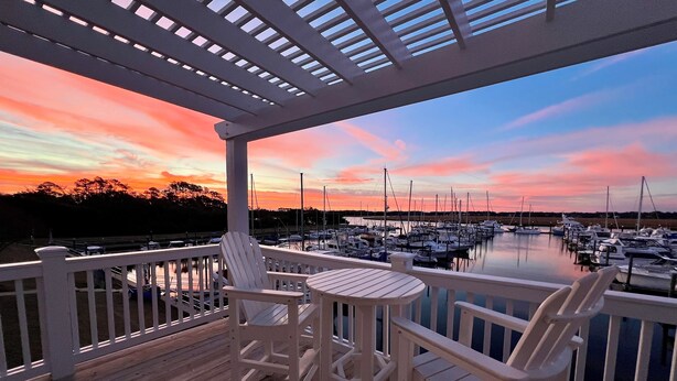 Eine Holzterrasse mit weißen Stühlen und einem Tisch, mit Blick auf einen Hafen voller Boote und einem Sonnenuntergang.
