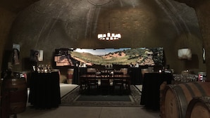 A rustic wine cellar with a large TV screen displaying a vineyard, wooden barrels, and a chandelier.