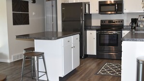 A modern kitchen with stainless steel appliances, white cabinetry, and a bar stool. - Next Door To Lambeau Field /Sleeps 10+ (Green Bay)