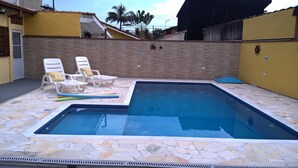 Une piscine aux bords recouverts de carrelage en pierre et une terrasse carrelée, le tout entouré d’un mur de briques et d’un bâtiment jaune.