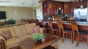 A modern kitchen with wooden cabinets, a stainless steel refrigerator, and a dining area with wooden chairs and a brown sofa. - Ocean Front Private Beach, Pool/Hot Tub, Pool Bar, Marina with Cafe (Tavernier)
