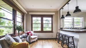 A kitchen with wooden floors, a bar area with stools, a window with a view of greenery, and a white chair with a throw blanket. - Eurunderee House - nestled in Mudgee’s famous vineyards (Eurunderee)