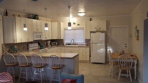 Une cuisine moderne avec des meubles blancs, un îlot central doté de tabourets, un réfrigérateur et une fenêtre avec des stores.