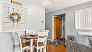A dining area with a wooden table and white chairs, a wall clock, and a sign above the door. - 'Escape Plan'  great location for year round fun (Mancelona)