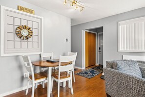 Un coin repas avec une table en bois et des chaises blanches, une horloge murale et une enseigne au-dessus de la porte.