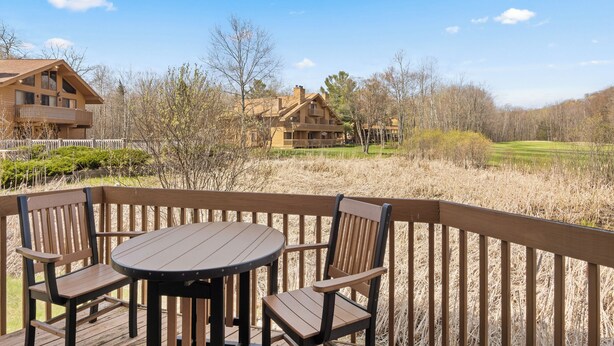A wooden deck with a round table and chairs, overlooking a building with a balcony and a golf course in the background. - 'Escape Plan'  great location for year round fun (Mancelona)