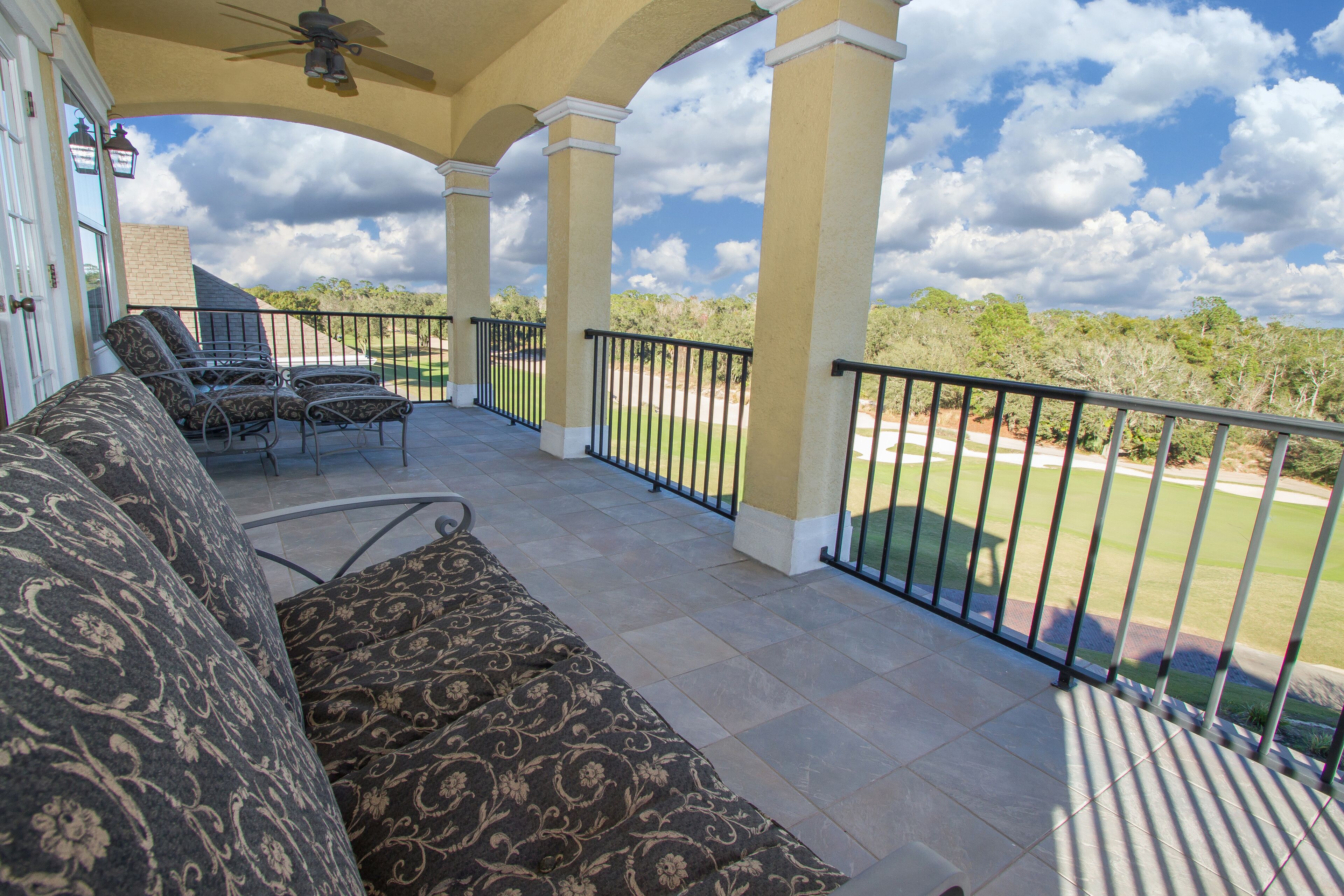 A covered patio with a view of a golf course, featuring a seating area with patterned cushions and a ceiling fan.