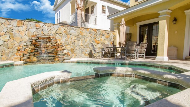 Ein Pool mit einem Wasserfall aus Stein, umgeben von einer Steinmauer, sowie eine Terrasse mit Stühlen und einem Tisch.